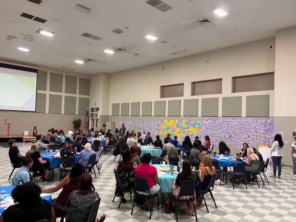 A large Butte County community gathering seated at round tables, facing a wall covered in hundreds of coloured sticky notes contributed during a group exercise.